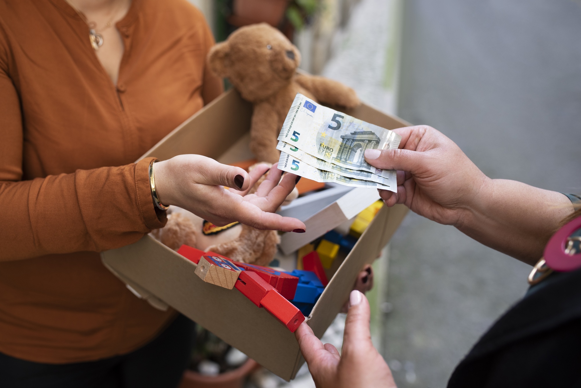 Das Bild zeigt einen Verkaufsvorgang in Großansicht, bei dem ein Mädchen mit umgehängtem Bauchladen ein Warenstück übergeben hat und jetzt von der Hand des Käufers im Gegenzug mehrere Geldscheine entgegen nimmt.