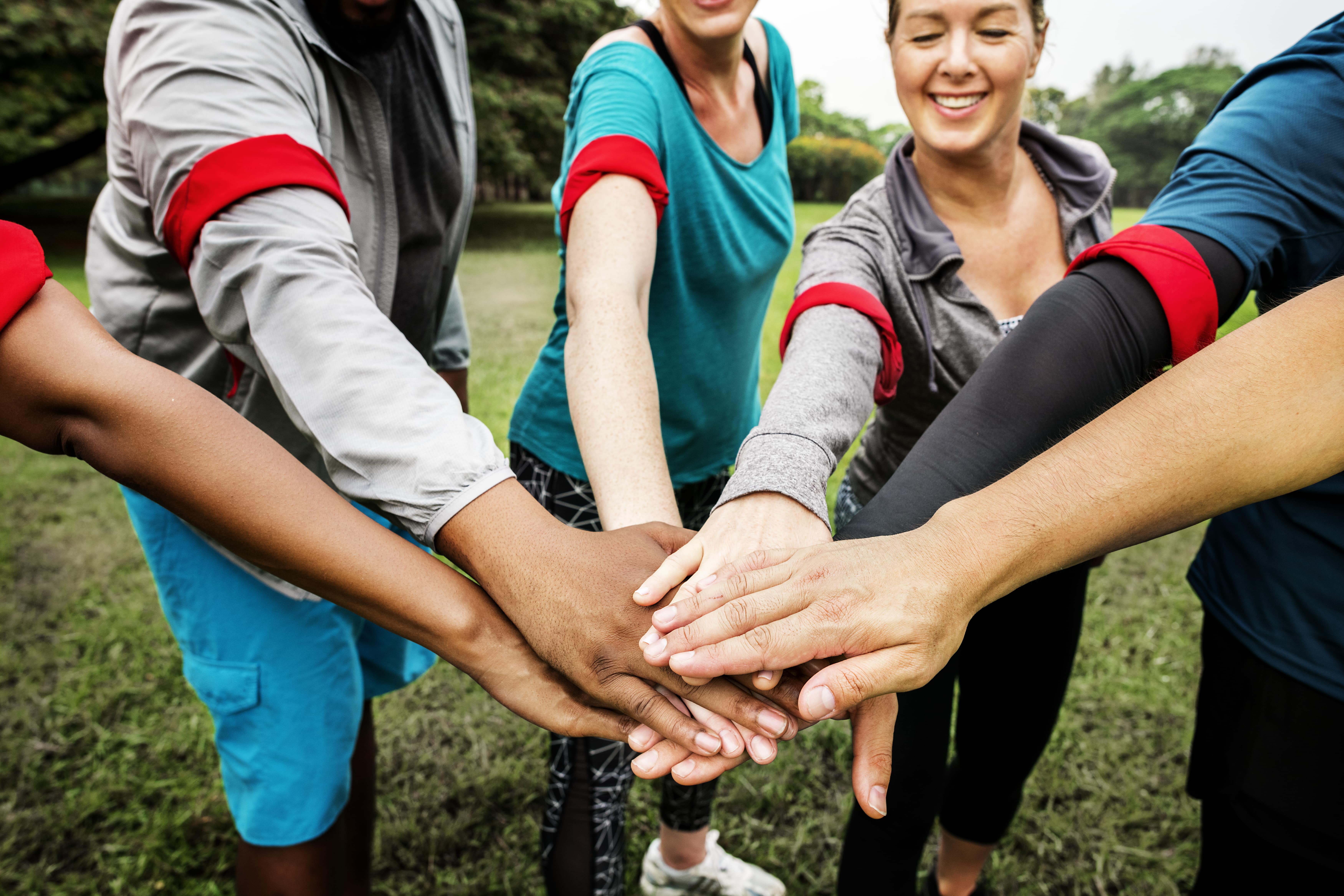 Das Bild zeigt eine Gruppe von Männern und Frauen in Sportbekleidung. Sie strecken jeweils einen Arm nach vorne und legen zum Zeichen des gemeinsamen Handelns die Hände aufeinander. b
